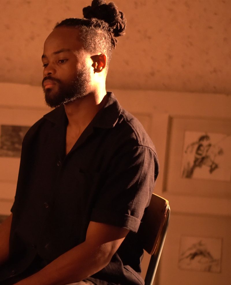 Zayane Abdou Mhoussine, seated in his studio in warm lamplight, framed drawings on the wall behind him.